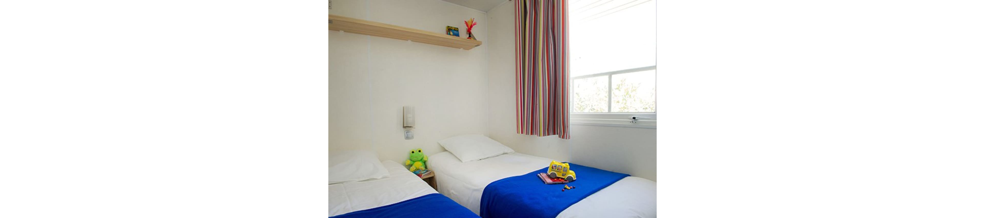 A small, bright bedroom features two single beds with white linens and vivid blue blankets. A window with striped curtains lets in natural light, illuminating the simple white walls. A green stuffed frog sits on one bed, while a toy bus and small figures rest on the other. A wooden shelf with a few items and a wall-mounted device add subtle details to the minimalist space.
