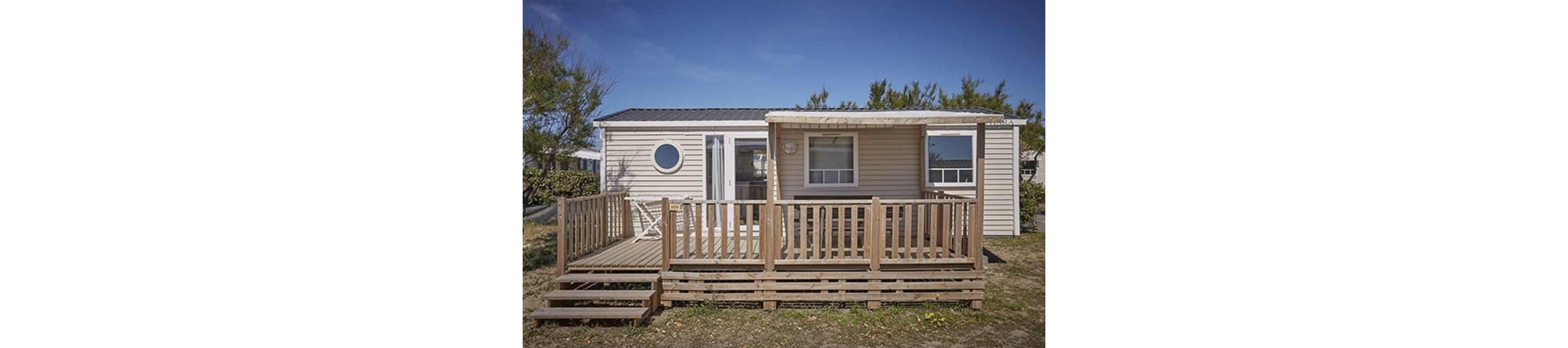 A compact beige mobile home with a wooden deck is shown under a clear blue sky. The structure features a front porch with railings, steps leading up from the ground, and large windows. A distinctive round window and sliding glass door add visual interest to the facade. Sparse grass and surrounding trees suggest a quiet, natural setting, possibly in a campground or holiday park.