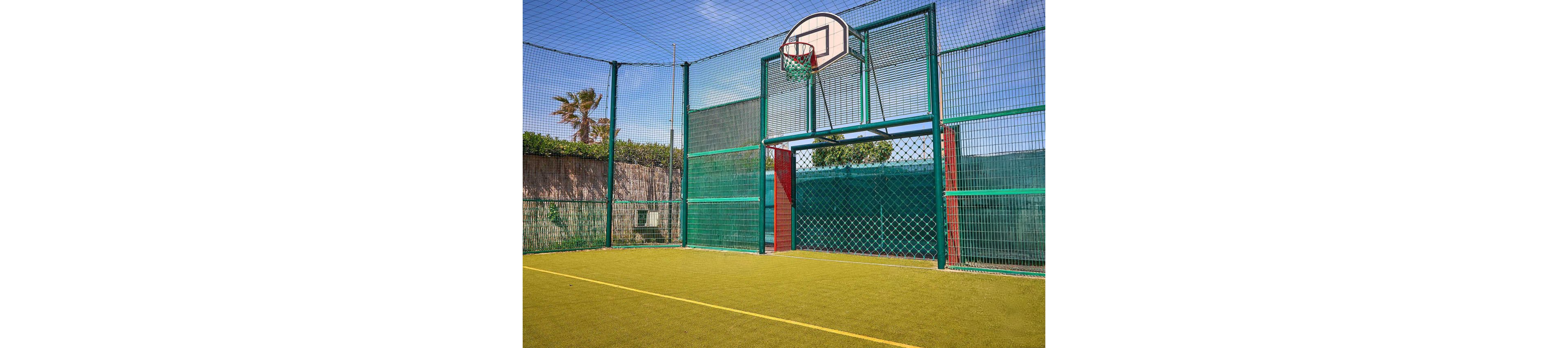 Two adults play a casual game of basketball on a fenced outdoor court. A woman in athletic wear throws a ball toward a man standing near the hoop. The court has bright green turf, a metal fence, and a clear blue sky with palm trees in the background. The scene conveys an active, sporty lifestyle and warm, sunny weather.
