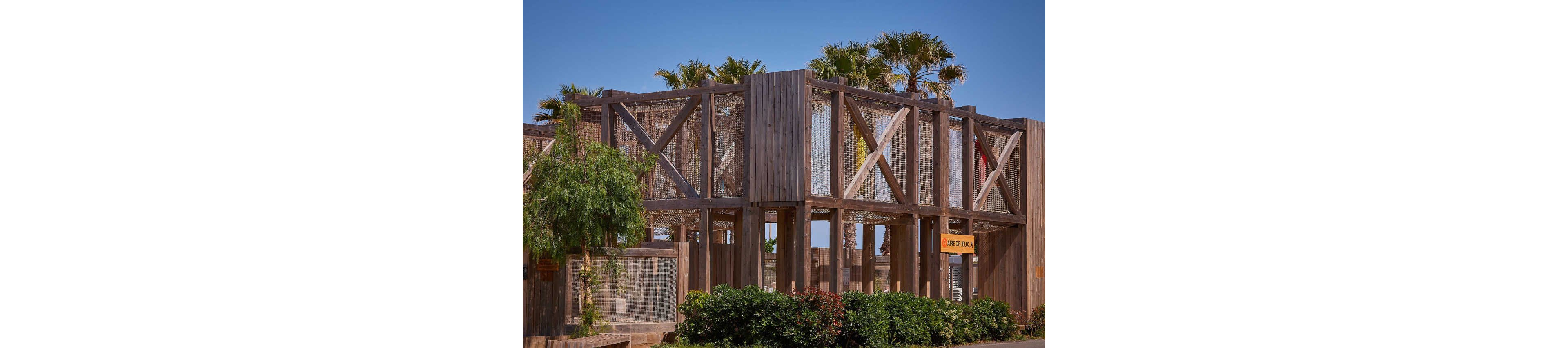 A large wooden aviary structure stands outdoors under a clear blue sky. The construction features tall vertical beams, cross-bracing, and mesh panels enclosing the space. Palm trees rise behind and above the structure, adding a tropical feel. Green shrubs and plants line the front, softening the geometric architecture. A small yellow sign is visible on the right side of the building.