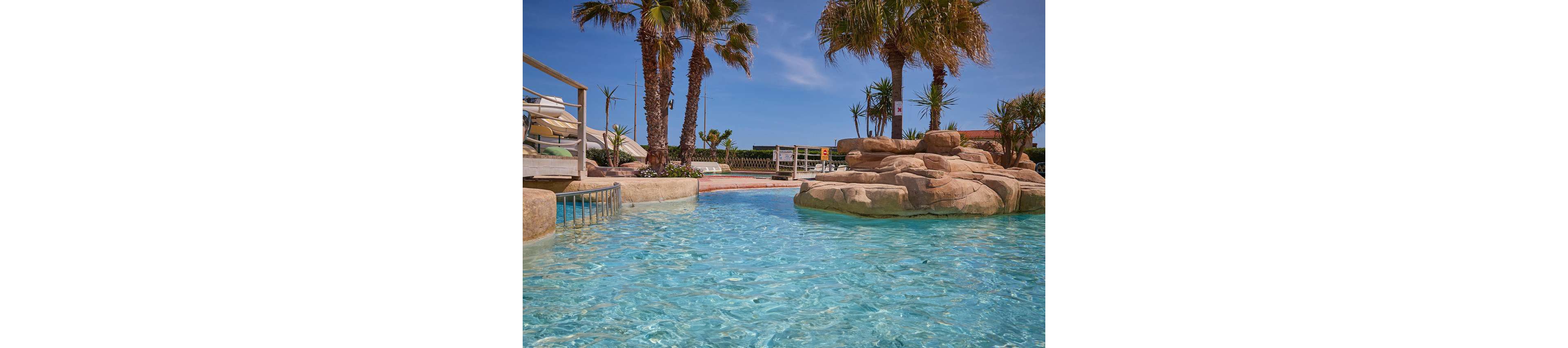 A woman and two young children play together in a shallow outdoor pool. The kids use inflatable toys, including a tire-shaped float and arm floaties, while the adult supervises nearby. Palm trees, rocks, and clear blue water create a tropical resort atmosphere. The bright sunlight and clear sky suggest a warm, relaxing vacation setting.