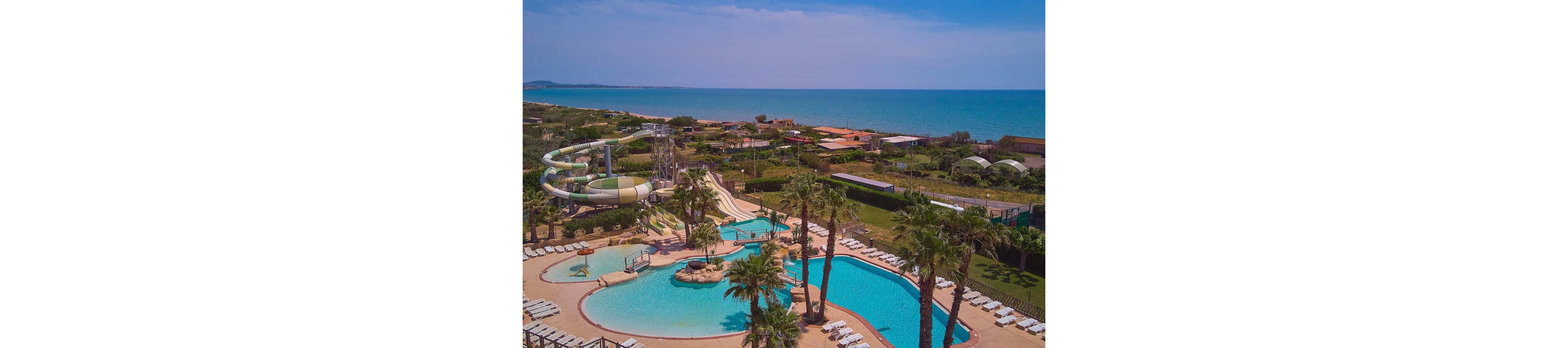 A large resort swimming pool complex is shown from above near a coastline. Multiple curving waterslides and splash areas are surrounded by palm trees and sun loungers. The bright blue pools contrast with the green landscaped gardens and the deep blue sea in the background. Buildings, sports courts, and pathways extend toward the beach under a clear, sunny sky.