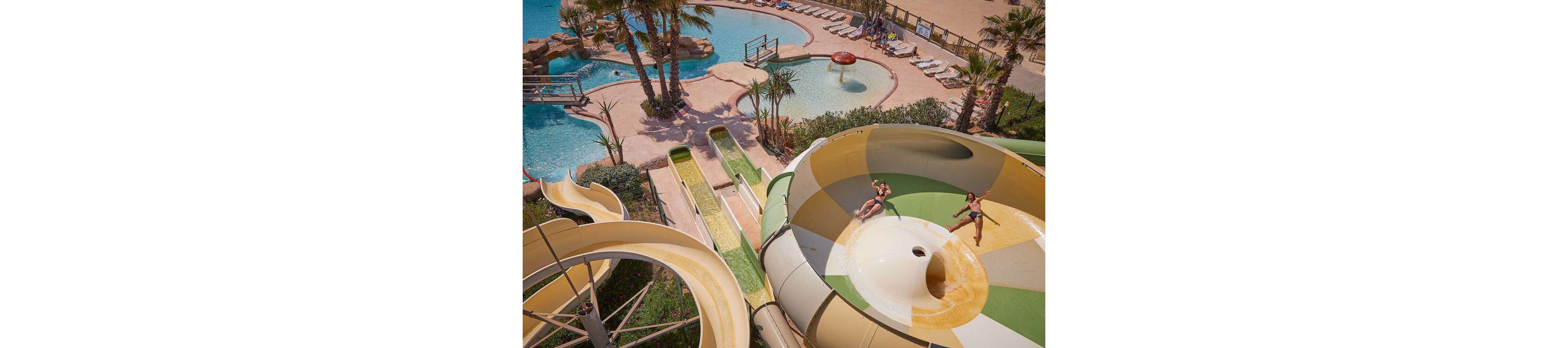 A group of people ride a large spiral water slide at an outdoor water park. The slide empties into a pool surrounded by palm trees, sun loungers, and additional pools. The scene is bright and sunny, with clear blue water and sandy-colored structures creating a tropical vacation atmosphere. The image conveys fun, leisure, and summertime recreation.