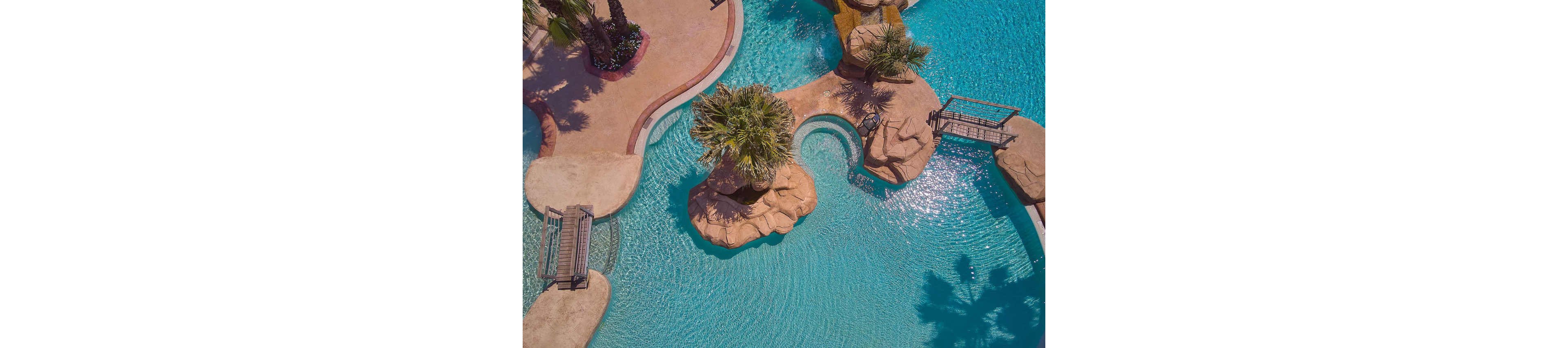 A turquoise resort swimming pool is seen from above, featuring curving edges and clear water. Palm trees grow from rocky islands in the middle of the pool, creating a tropical oasis feel. Small bridges connect the poolside walkways to the central rock formations. The scene suggests a relaxing vacation setting under bright sunlight.