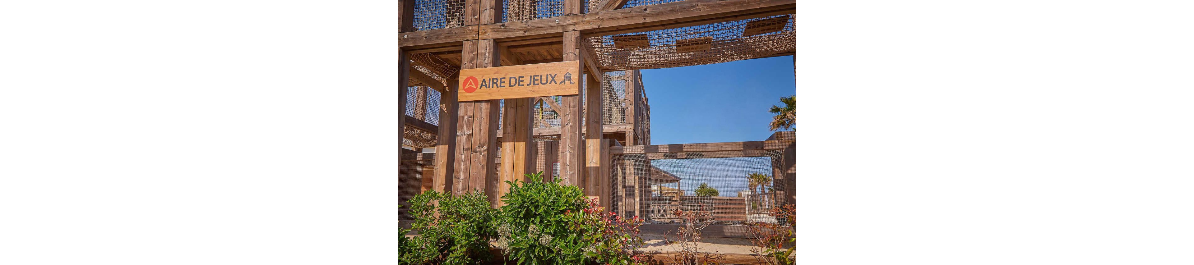 A young child is captured mid-jump in front of a large wooden playground structure. The scene takes place outdoors on a sunny day, with blue sky and palm trees visible in the background. A sign reading "AIRE DE JEUX" is mounted on the structure, indicating a play area. Colorful plants and flowers are in the foreground, adding a natural touch to the recreational setting.
