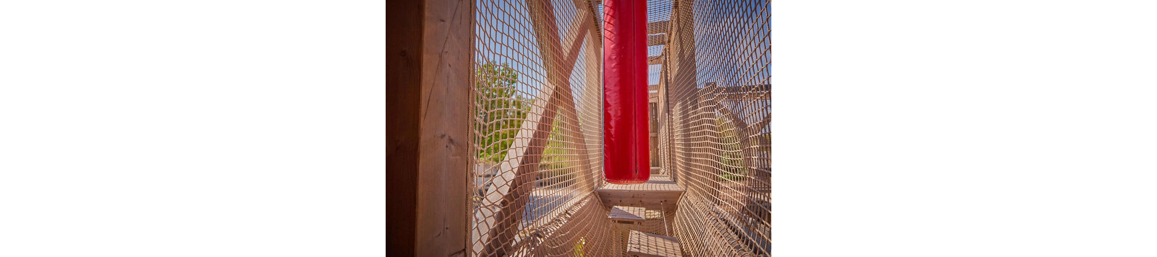 Two children play on a suspended rope bridge at an outdoor playground. The older child in a gray shirt and blue shorts leans against the netting while moving forward. A younger child in a floral dress stands further back on the wooden planks. Bright sunlight and warm tones create a fun, adventurous atmosphere. Wooden structures and netted sides emphasize the climbing and balancing activity.