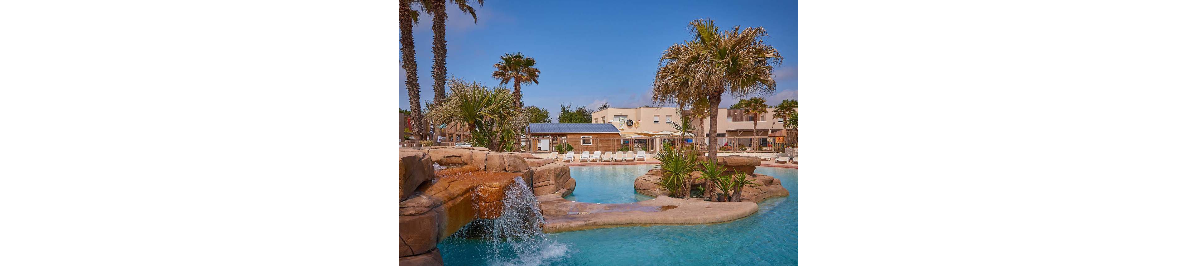 A large resort-style swimming pool features a rock waterfall cascading into clear blue water. Palm trees and tropical plants surround the pool, creating an inviting vacation atmosphere. In the background, sun loungers are lined up in front of low-rise buildings and a poolside bar or café. The scene suggests a sunny, relaxing day at a holiday resort.