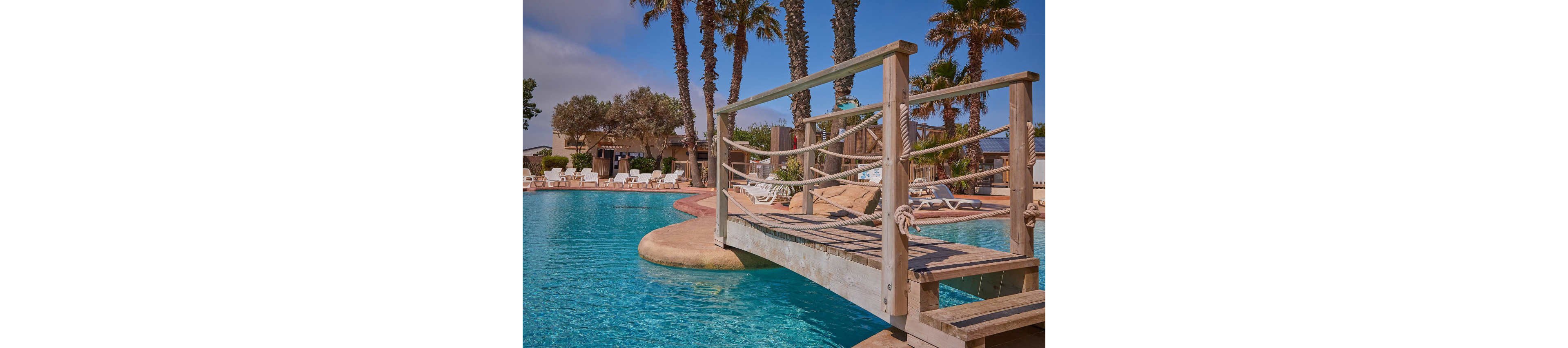 A resort-style swimming pool features a small wooden bridge with rope railings crossing over the water. Sun loungers and umbrellas line the poolside deck, suggesting a relaxing vacation setting. Tall palm trees and other greenery surround the area, adding a tropical atmosphere. Buildings in the background indicate a hotel or holiday complex under a clear, sunny sky.