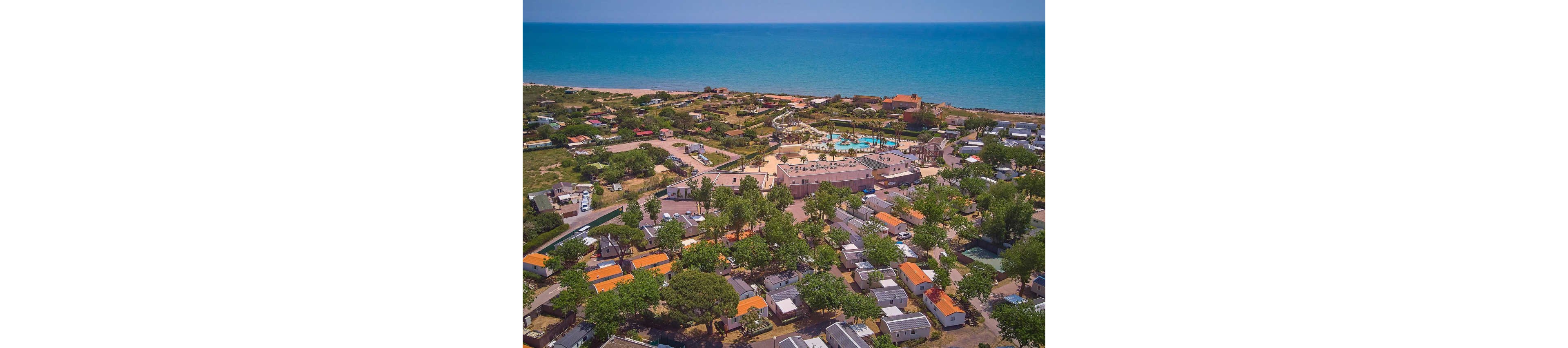 A seaside holiday resort is captured from above, showing rows of mobile homes and cabins surrounded by trees. In the center of the complex, a large swimming pool area with sun loungers and palm trees stands out. The resort is situated directly along a wide sandy beach that meets a calm blue sea. Roads and pathways connect the various accommodations, creating an organized vacation village layout.