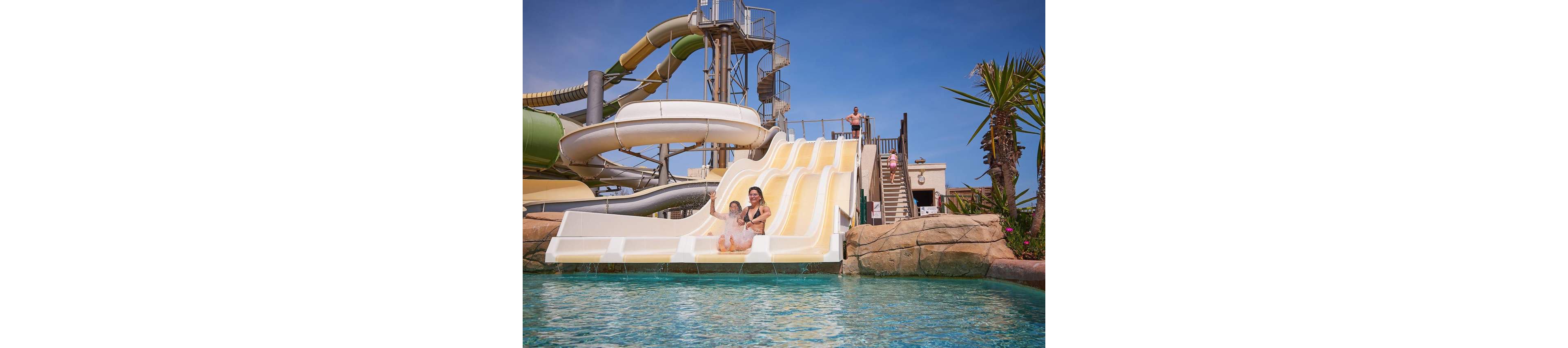 A woman and a young child slide down a wide multi-lane water slide into a clear blue pool. The scene is set in an outdoor water park with additional twisting tube slides and metal stairways in the background. Other visitors stand on the stairs and at the top platform waiting their turn. Palm trees and rock-like structures frame the attraction under a bright, sunny sky, creating a fun vacation atmosphere.