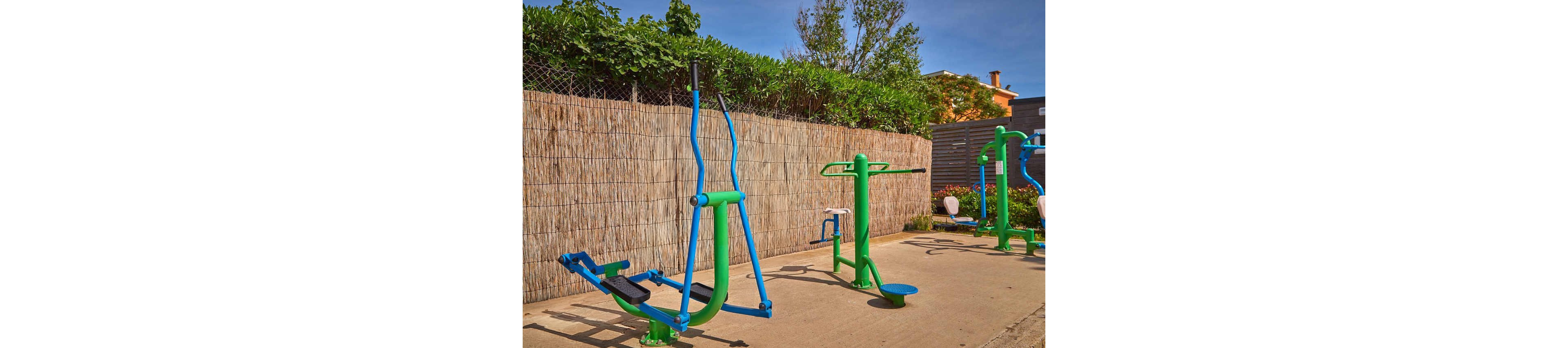 Two adults work out on brightly colored outdoor fitness equipment in a sunny recreational area. The woman uses an elliptical-style machine while the man stands on a balance platform, both dressed in athletic clothing. The setting features a paved surface, green metal machines, and a tall fence with lush greenery behind it. Clear blue sky and warm light suggest a pleasant, active day outside.