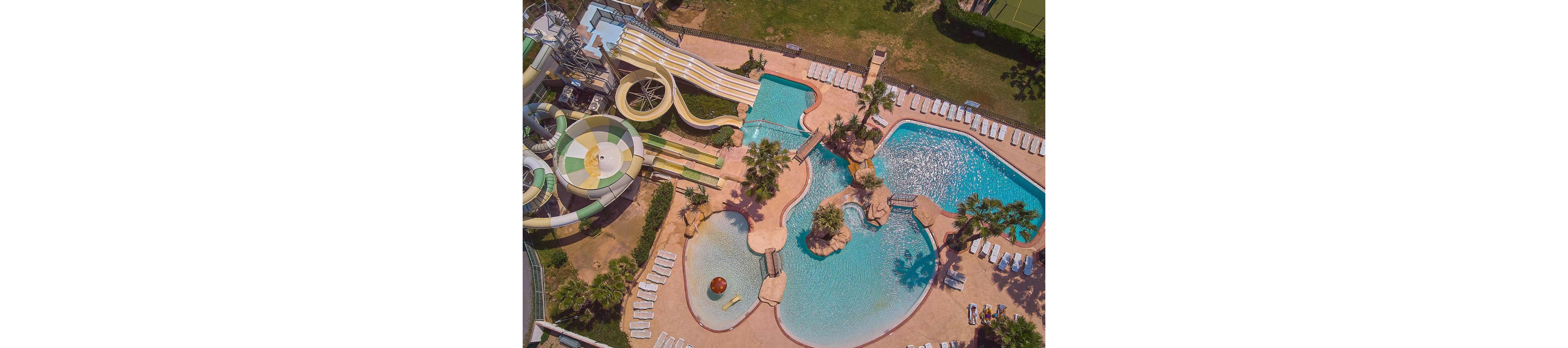 A large resort-style water park is captured from above, showcasing multiple interconnected swimming pools and winding water slides. Curved slides in shades of yellow and green descend into the pool area, surrounded by palm trees and landscaped rock features. Numerous white lounge chairs line the pool decks, suggesting a relaxing vacation atmosphere. The bright sunlight and clear blue water create a warm, inviting summer scene.