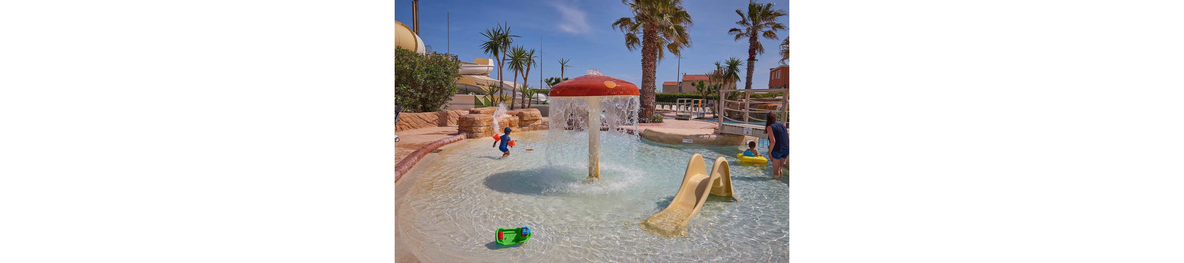 Young children play in a shallow splash pool at an outdoor water park. A large mushroom-shaped fountain sprays water while a small yellow slide leads into the pool. Palm trees, waterslides, and resort buildings are visible in the background under a clear blue sky. Colorful pool toys float in the water, creating a fun and family-friendly vacation atmosphere.