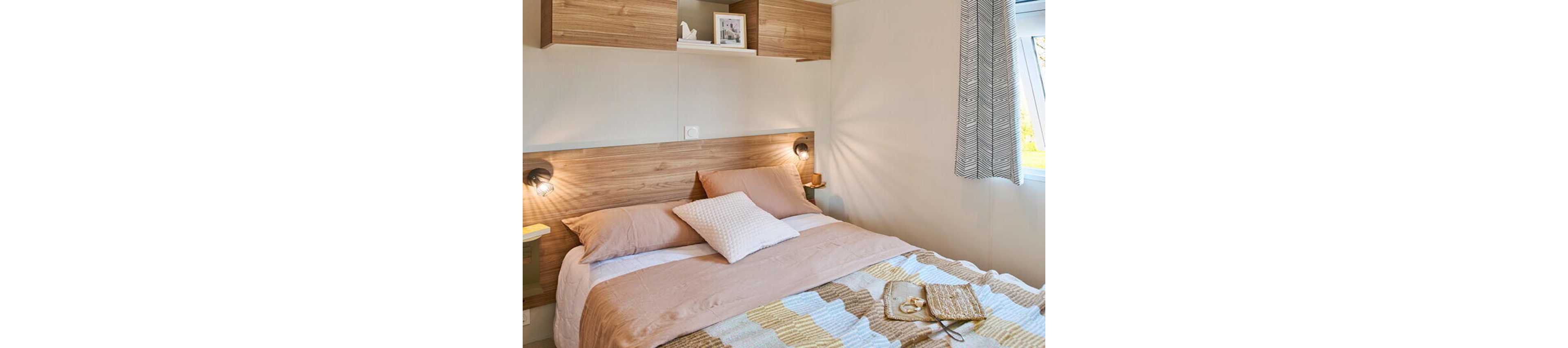 A neatly made double bed with neutral-toned bedding is the focal point of this modern bedroom. Wooden headboard panels and overhead storage cabinets add warmth and functionality to the space. Soft wall-mounted lights create a cozy ambiance, while a window with patterned curtains lets in natural daylight. A tray with accessories and a book on the bed adds a relaxed, lived-in touch.