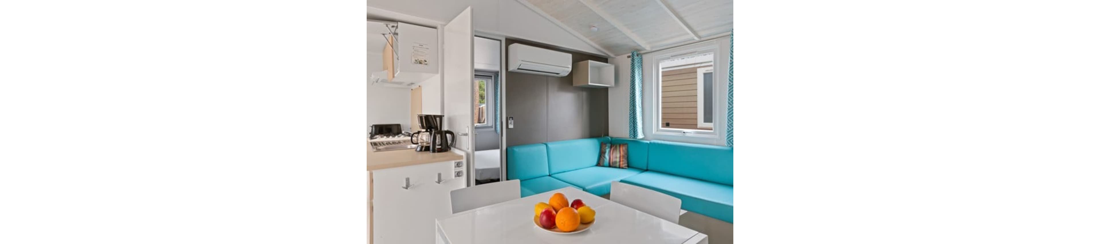 Compact modern living space with a turquoise corner sofa and white dining table. A bowl of assorted fresh fruit sits on the table in the foreground. The room includes a small kitchenette with a coffee maker and stove on the left. Large windows with patterned curtains and a wall-mounted air conditioner create a bright, comfortable vacation cabin atmosphere.
