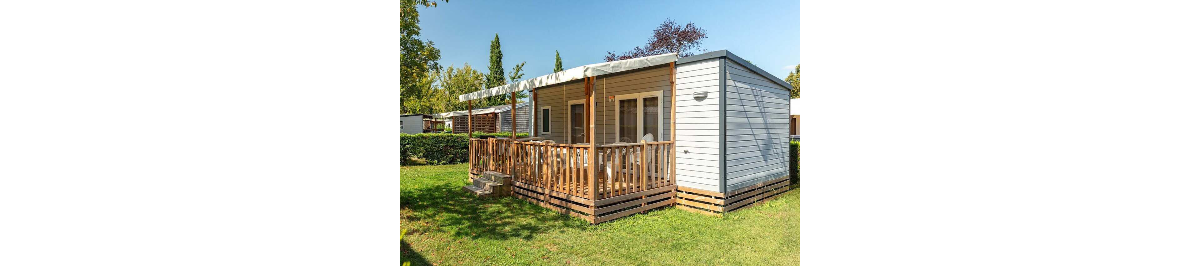 A modern mobile home with light gray siding features a spacious wooden terrace and railing. White outdoor chairs sit under a covered porch, suggesting a comfortable place to relax. The home is surrounded by green grass, hedges, and tall trees under a clear blue sky. Other similar units are visible in the background, indicating a holiday park or campground setting.