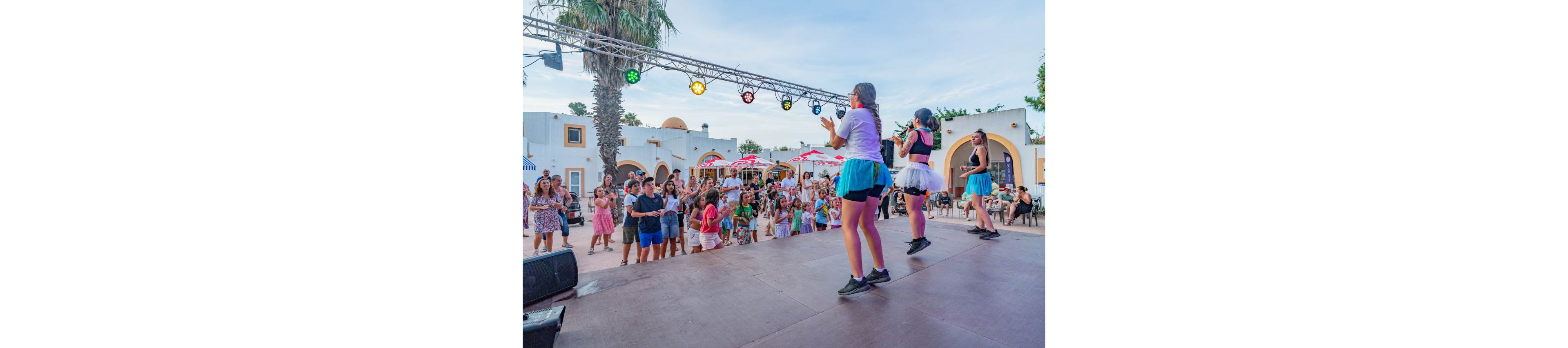 Three performers in colorful outfits entertain a crowd from an outdoor stage at a resort. The audience, including families with children, stands in an open courtyard surrounded by white buildings and palm trees. Stage lights hang overhead, adding a festive atmosphere to the early evening scene. The image conveys a lively vacation entertainment event with people clapping and watching the show.