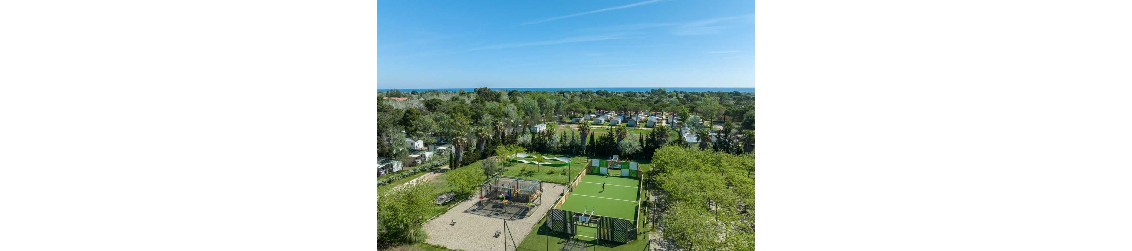 An aerial view shows a green holiday campsite near the sea, with rows of mobile homes and caravans surrounded by trees. In the foreground, there is a fenced sports court where a person is playing on the artificial turf. Adjacent to the court is a small playground area with equipment. The background reveals dense greenery leading to a blue coastline under a clear sky, creating a relaxed vacation atmosphere.