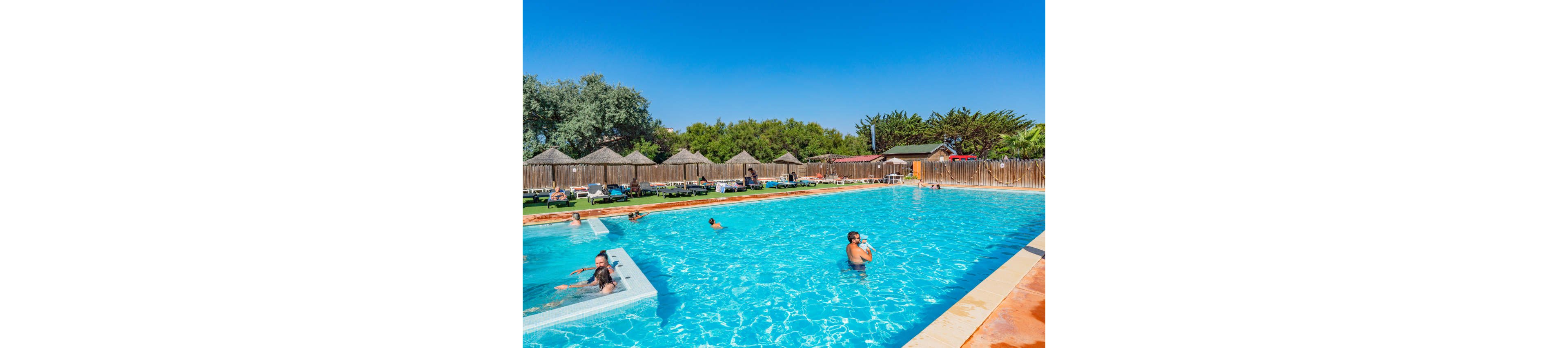 An outdoor swimming pool is filled with people enjoying a sunny day at a resort. Adults and children swim and wade in the bright blue water, while others relax on loungers around the pool. Thatched-roof cabanas and a wooden fence line the background, framed by green trees and clear blue sky. The scene conveys a warm, leisurely holiday atmosphere in a well-maintained pool area.