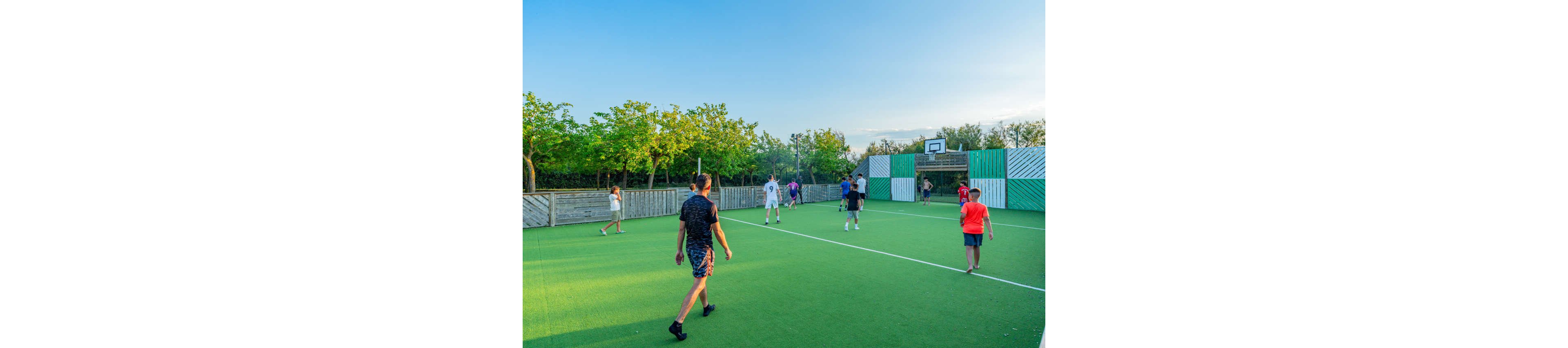A group of children and teenagers play soccer on a bright green outdoor sports court. The scene is set in a fenced recreational area with trees and greenery surrounding the field. A basketball hoop and striped panels are visible in the background, indicating a multi-sport facility. The warm evening light and clear sky create a relaxed, active atmosphere.