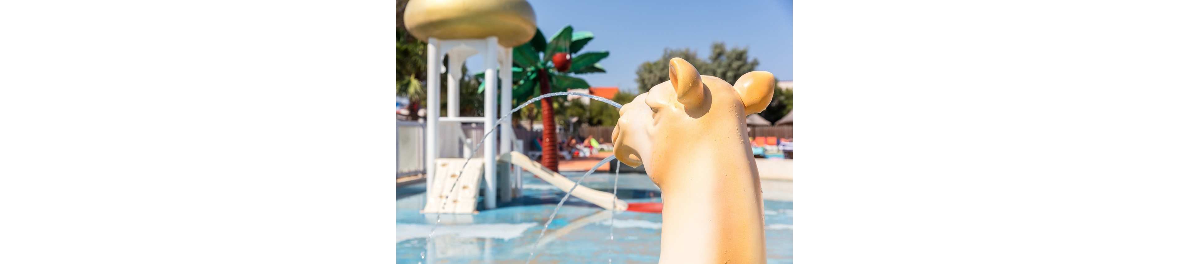 A camel-shaped fountain statue sprays water into a shallow outdoor pool. In the background, a small slide structure and a large artificial palm tree add to the playful water park setting. Bright sunlight and clear skies create a warm, summery atmosphere. The scene suggests a family-friendly recreational area designed for children.