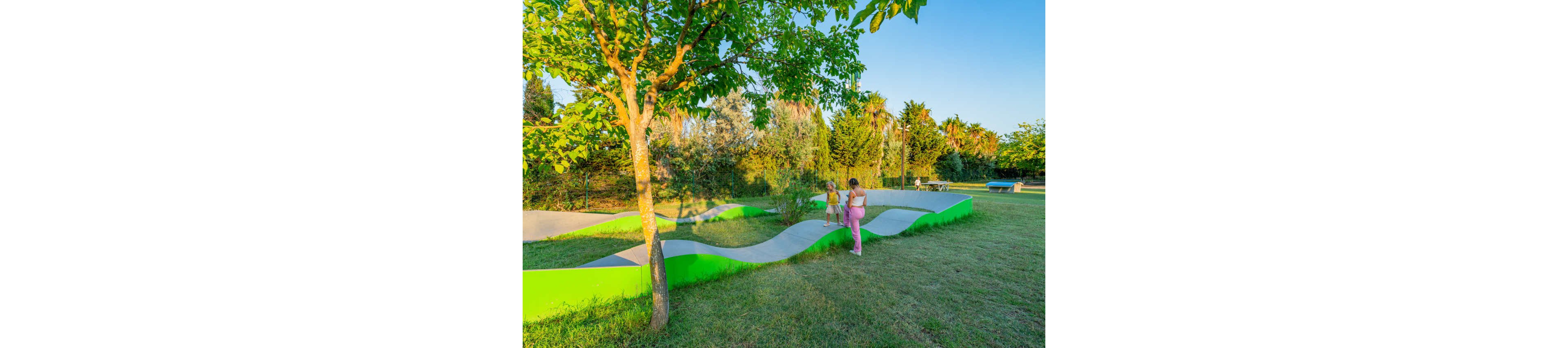 Two young children walk along a wavy concrete pathway in a sunny park. The path is bordered by bright green edges that contrast with the surrounding grass. Trees and shrubs provide a lush, natural backdrop under a clear blue sky. The warm evening light creates a relaxed, playful outdoor atmosphere.