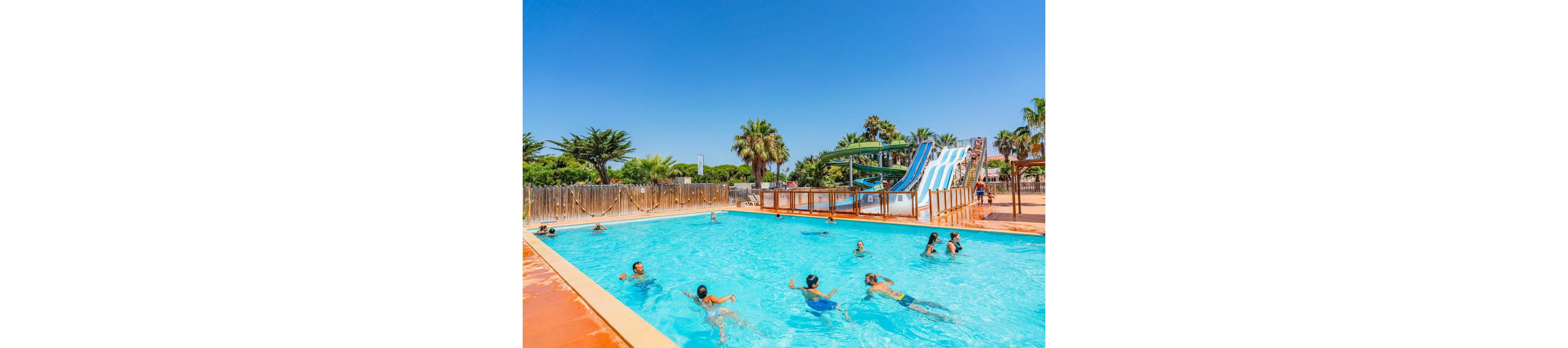 An outdoor swimming pool is filled with people swimming and playing in the water. In the background, several colorful water slides rise above the pool area, suggesting a fun water park setting. Palm trees and clear blue sky create a bright, sunny, vacation atmosphere. The scene conveys leisure, summer holidays, and family-friendly recreation.