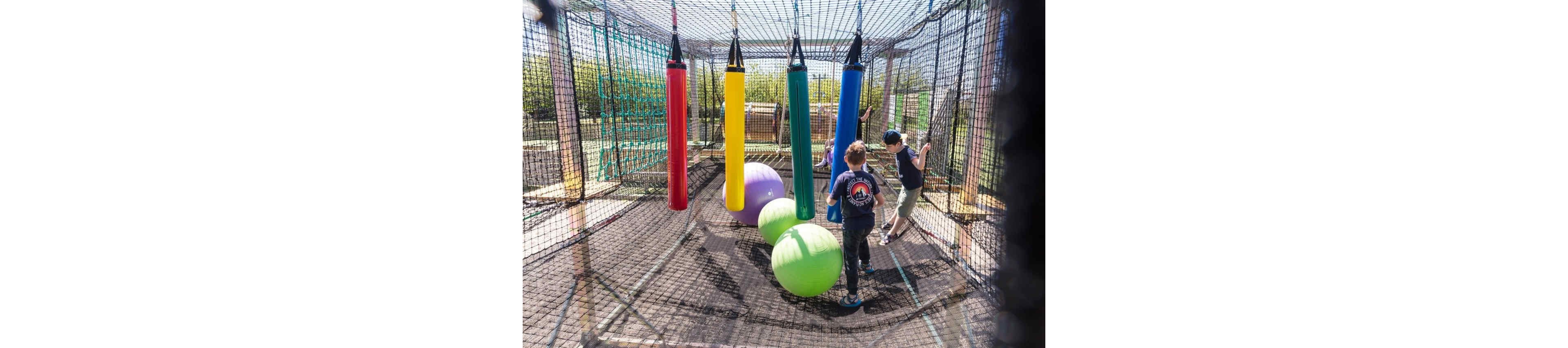 Children play together in an outdoor obstacle course enclosed by safety netting. Large hanging padded cylinders in red, yellow, green, and blue create a playful challenge. Bright exercise balls rest on the ground as kids navigate through the structure. The sunny day and vivid colors give the scene an energetic, fun atmosphere.