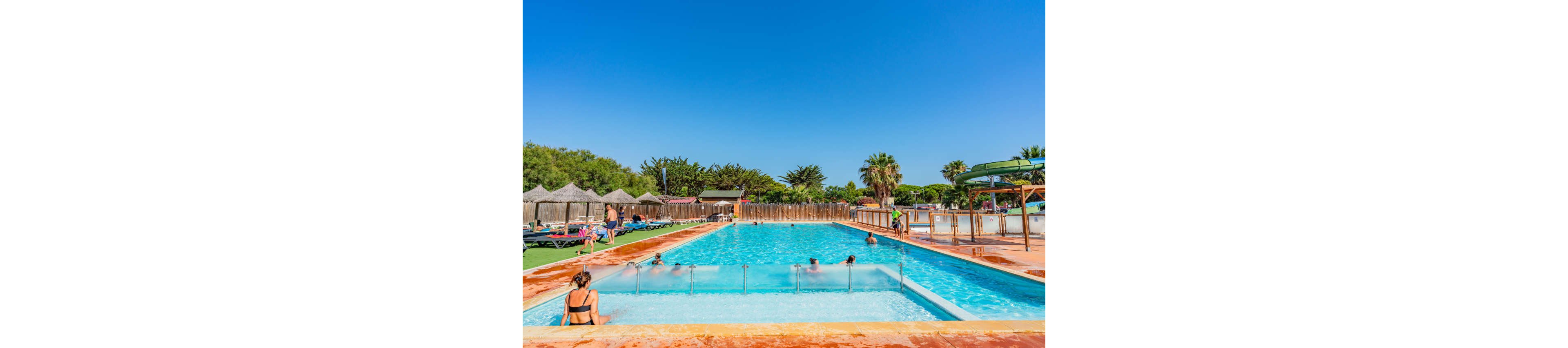 An outdoor swimming pool is filled with people relaxing and playing in the water under a clear blue sky. Sunbathers lounge on chairs and grass areas around the pool, some under thatched umbrellas. Trees and wooden fencing surround the pool area, giving it a resort-like atmosphere. The bright sunlight and vivid colors create a cheerful, summery vacation mood.