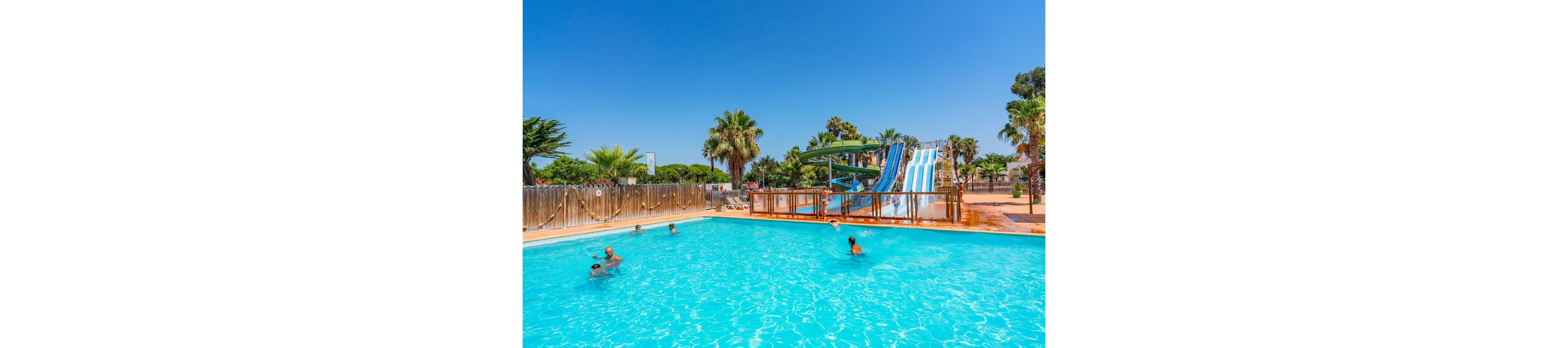 An outdoor swimming pool sparkles under a clear blue sky at a resort. Several people relax and play in the water, enjoying the sunny weather. Tall palm trees and other greenery surround the pool area, creating a tropical atmosphere. In the background, multiple large waterslides rise above the deck, adding a sense of fun and adventure.