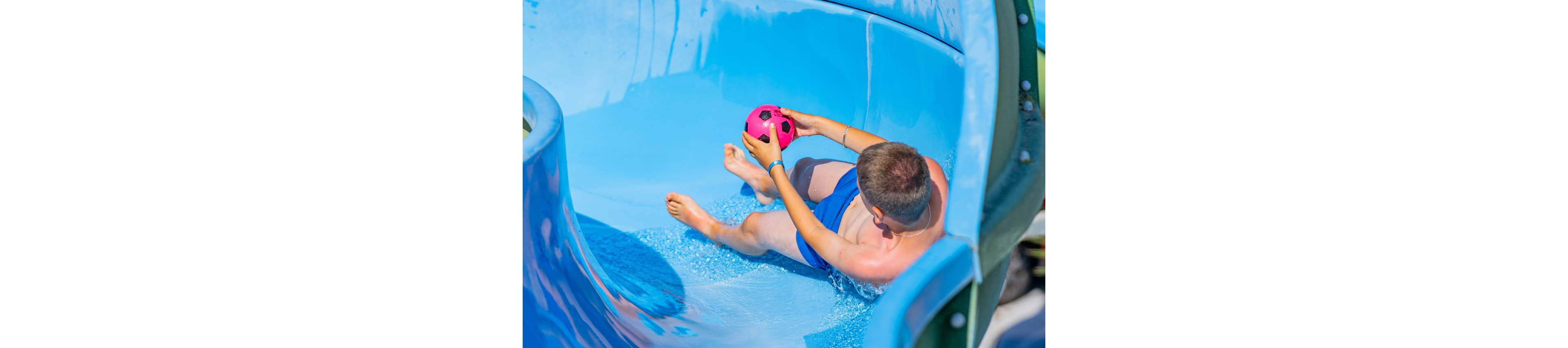 A young boy rides down a bright blue water slide while holding a pink soccer ball. He is seated in the flowing water, wearing blue swim trunks. Sunlight reflects off the wet slide surface, emphasizing the fun summer atmosphere. The scene captures playful activity at a water park or pool area.