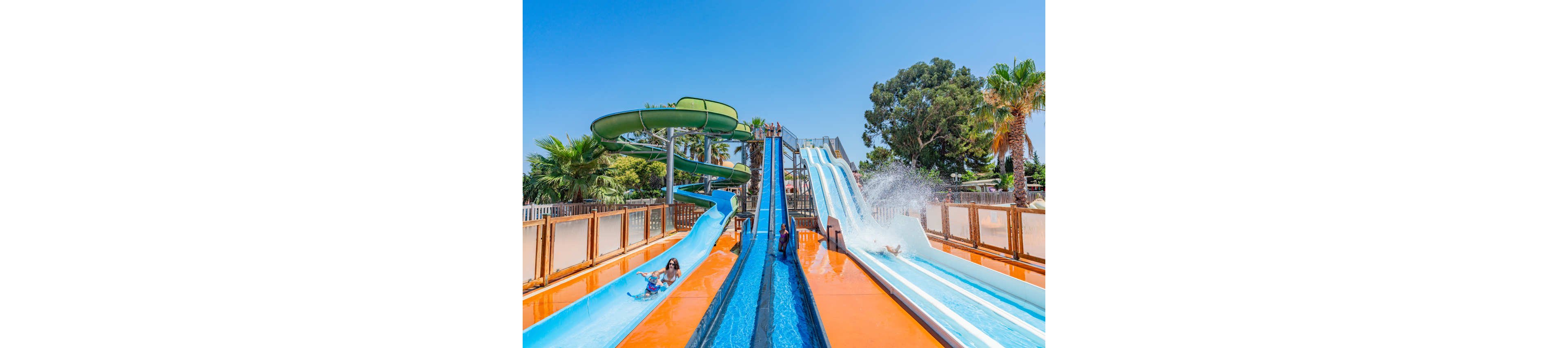 Several people slide down bright blue and white water slides at an outdoor water park. The scene shows multiple parallel slides and a large green tube slide winding in the background. Palm trees and lush greenery surround the attraction under a clear blue sky. Splashes of water and vivid orange flooring create a lively, energetic summer atmosphere.
