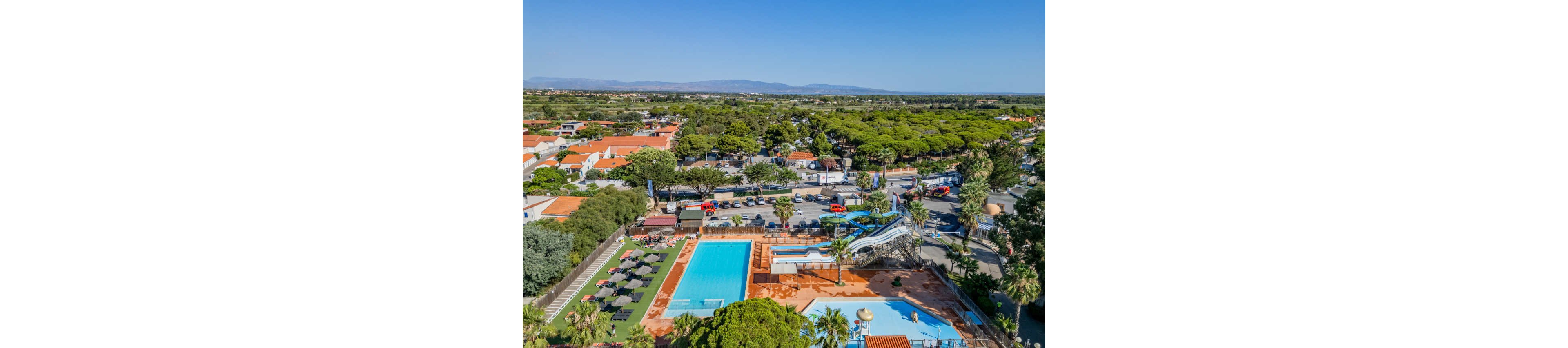 A large outdoor resort complex features multiple swimming pools and winding waterslides surrounded by palm trees. Sun loungers and umbrellas line the pool deck, while nearby buildings with red-tiled roofs indicate a holiday village setting. Beyond the resort, a green forested area stretches toward distant mountains under a clear blue sky. The bright sunlight and vivid colors create a relaxed vacation atmosphere.