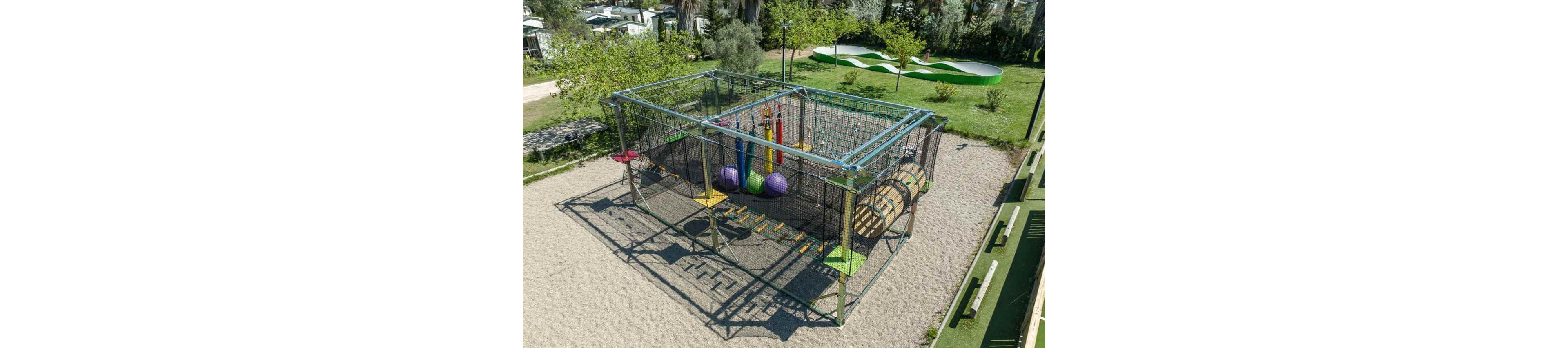 An elevated rope climbing playground structure stands in a sandy outdoor area. The cube-shaped frame is filled with nets, hanging obstacles, and colorful padded elements such as cylinders and balls. A wooden tunnel and balance steps add to the adventure course feel. Surrounding the structure are green lawns, trees, and nearby recreational areas, suggesting a family-friendly park or campsite setting.