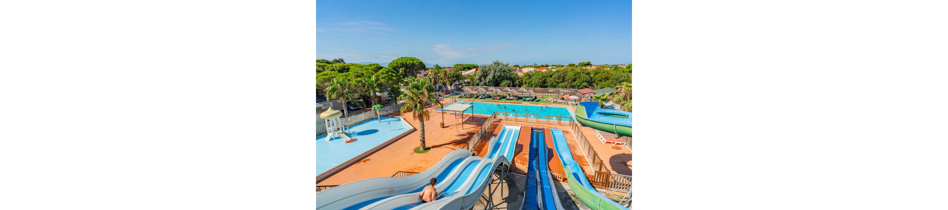 An outdoor water park features multiple large water slides leading down to a splash area. In the background, a spacious swimming pool is filled with people enjoying the sunny day. Palm trees, lounge chairs, and parasols surround the pool, creating a relaxed vacation atmosphere. The clear blue sky and bright colors emphasize a warm, family-friendly summer setting.