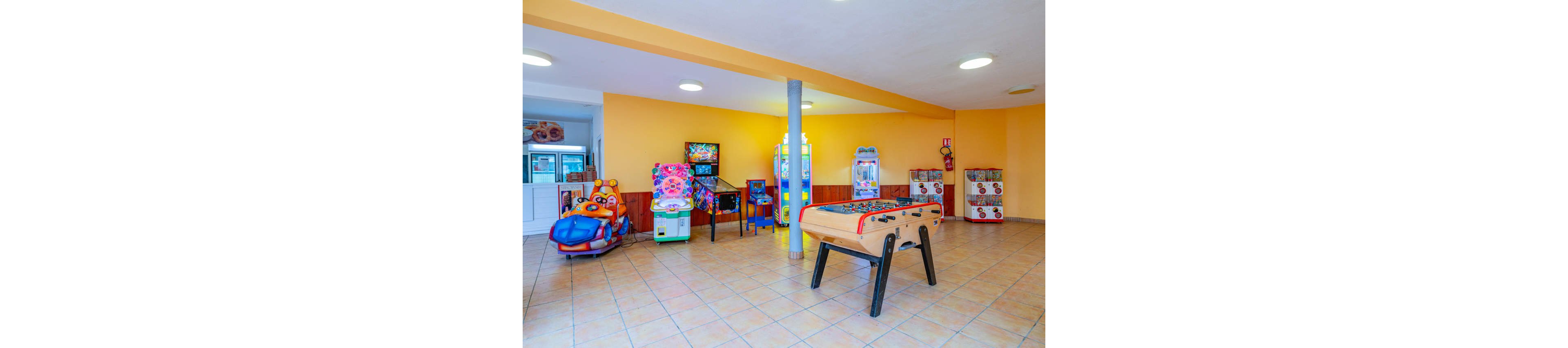 Colorful arcade machines and a foosball table fill a bright indoor recreation room. The space features tiled floors, yellow walls, and ceiling lights creating an inviting atmosphere. Various kiddie rides and game cabinets line the walls, offering entertainment for children and families. The room appears clean, spacious, and ready for playtime activities.