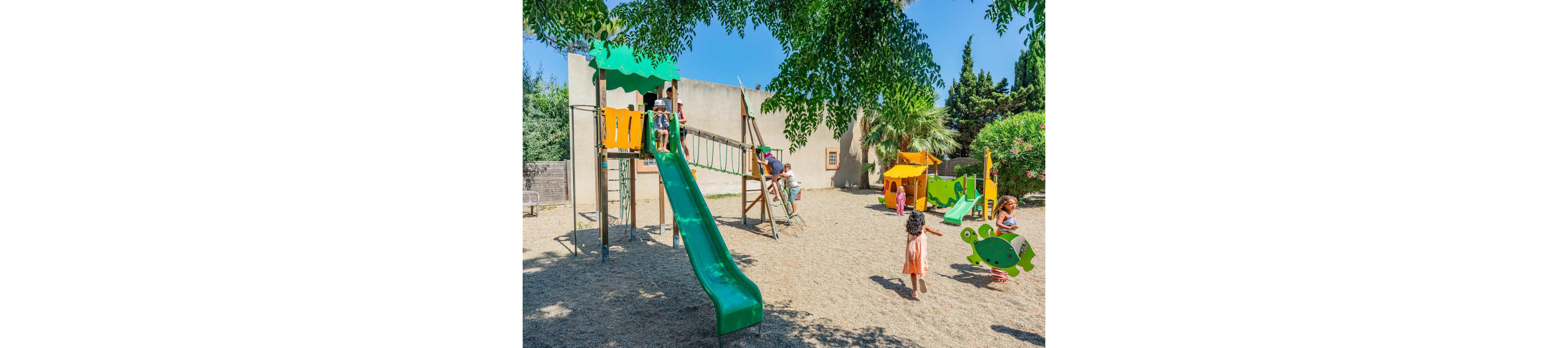Several young children play together in a sunny outdoor playground. A large green slide and climbing structure are the main focus, with kids climbing and sliding. Additional play equipment, including spring riders and smaller structures, is scattered around the sandy ground. Trees and shrubs provide greenery and shade, creating a cheerful and relaxed atmosphere.