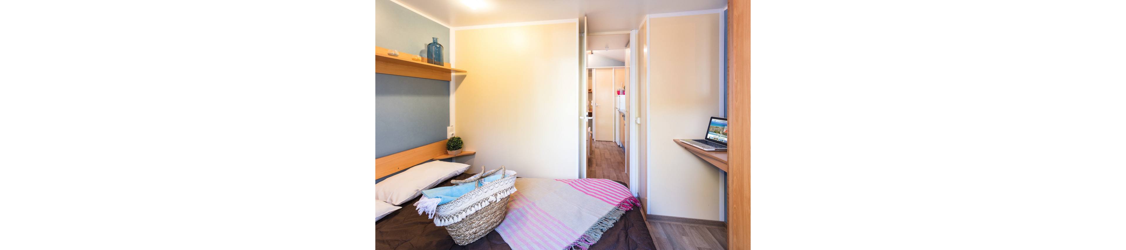 A compact bedroom features a neatly made bed with a striped throw and a woven basket placed on top. Light-colored walls and wood flooring create a bright, minimalist atmosphere. A narrow hallway leads to an adjacent room, adding depth to the space. A laptop sits on a small wall-mounted desk, suggesting a simple home office or study area within the bedroom.