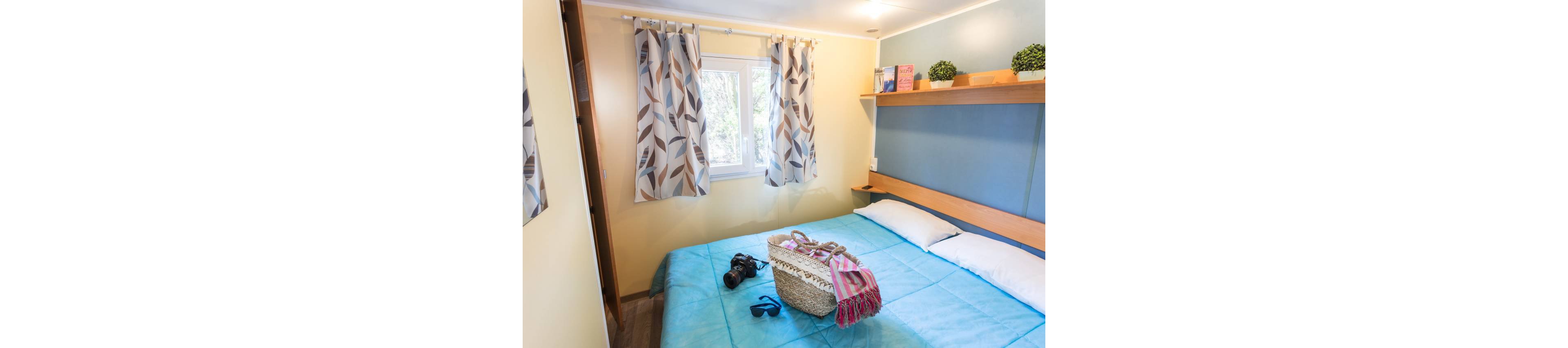 A small, bright bedroom features a neatly made double bed with a light blue bedspread. A woven beach bag, camera, and sunglasses rest on the bed, suggesting a holiday or weekend getaway. The room includes a window with patterned curtains, a wooden shelf with books and small potted plants, and soft ambient lighting. The overall atmosphere is relaxed and inviting, ideal for a vacation rental or guest room.