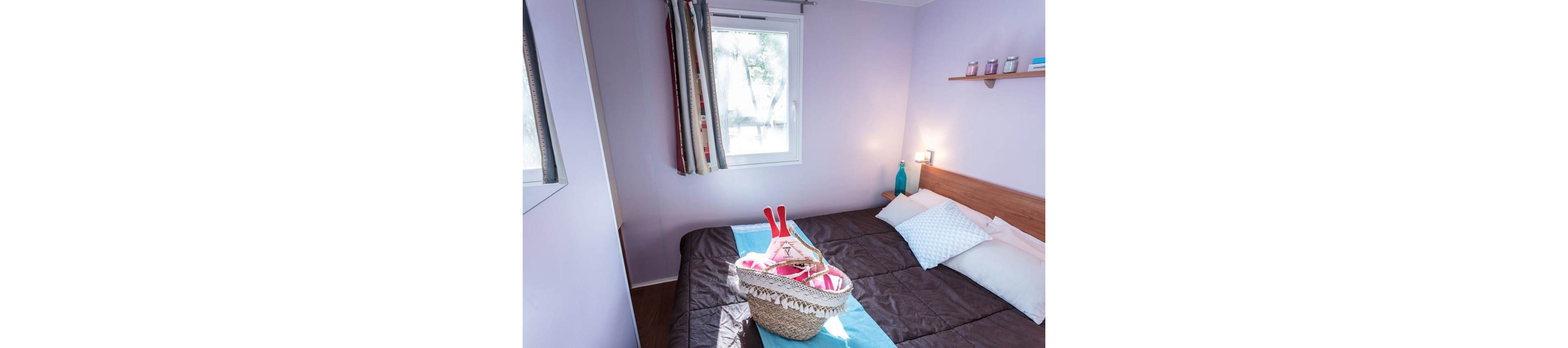 Compact bedroom with a double bed is decorated in soft pastel purple tones. A woven straw basket filled with beach items sits on the neatly made bed with a blue runner. Natural light enters through a small window with patterned curtains, brightening the room. A wooden shelf with small jars and a turquoise vase-style lamp add simple decorative touches.