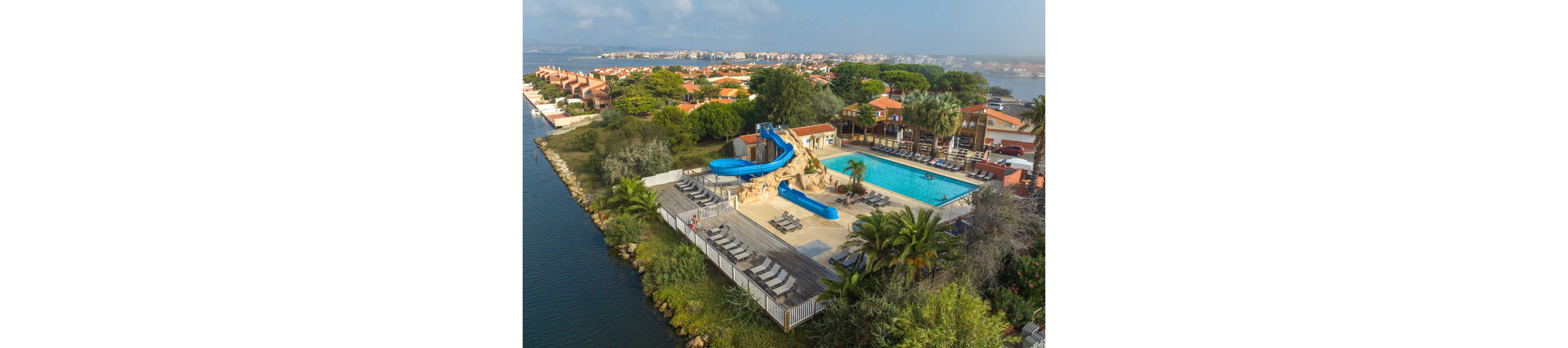An aerial view shows a coastal resort featuring an outdoor swimming pool and bright blue water slides built around artificial rocks. Sun loungers are neatly arranged on wooden decking beside the pool, surrounded by palm trees and lush greenery. The resort buildings with terracotta roofs extend into the background, bordered by calm water on one side and a distant shoreline community. The scene suggests a relaxed vacation atmosphere in a warm, seaside destination.