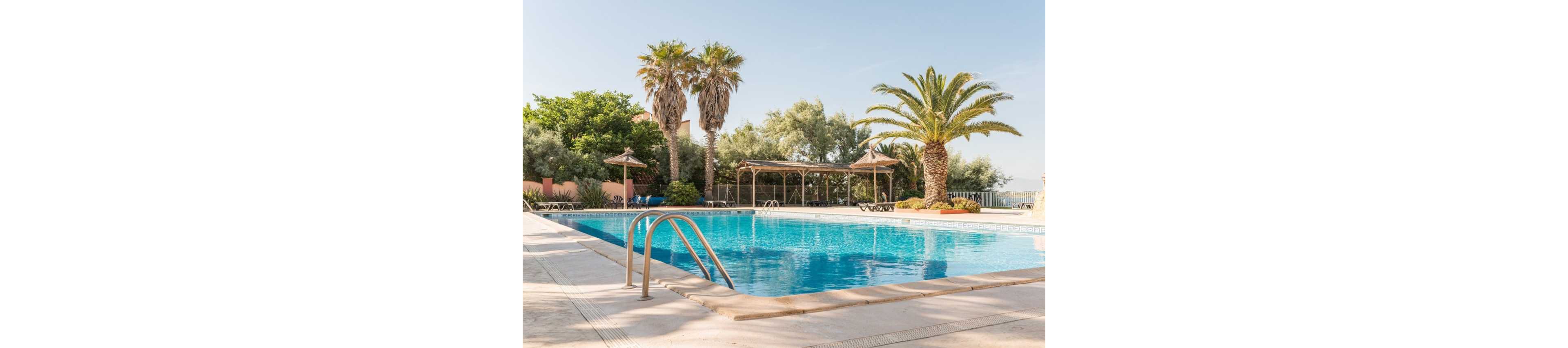 An inviting outdoor swimming pool glistens under clear blue skies at a resort. Palm trees and lush greenery surround the pool, creating a tropical atmosphere. Sun loungers and shaded pergolas line the poolside deck, suggesting a relaxing vacation setting. The calm water and empty chairs evoke a peaceful, tranquil mood.