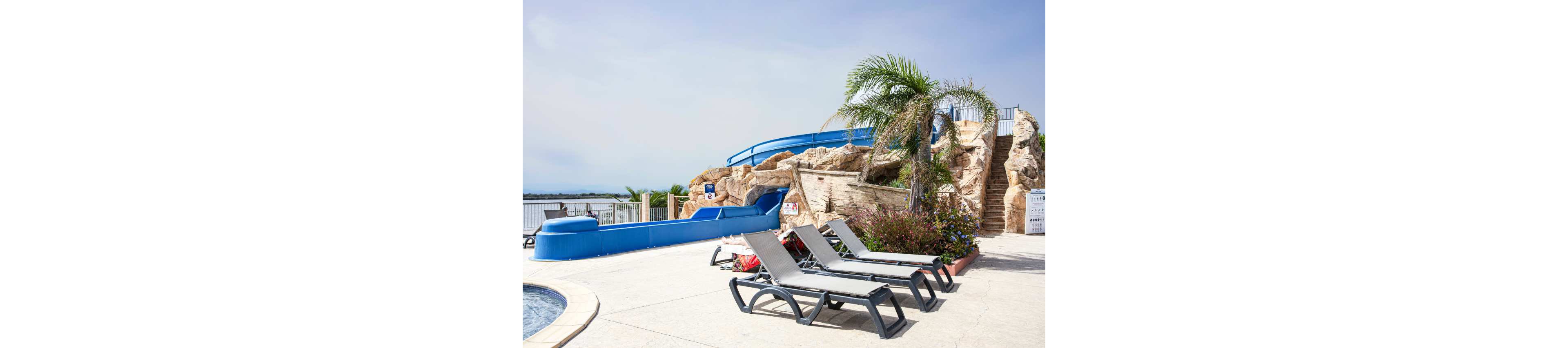 An outdoor pool area features several empty lounge chairs arranged on a sunlit deck. A large blue waterslide curves around artificial rock formations, ending near the pool. Palm trees and flowering shrubs add a tropical feel to the scene. A metal fence and distant water view suggest the pool is located near a lake or coastal area under a clear sky.