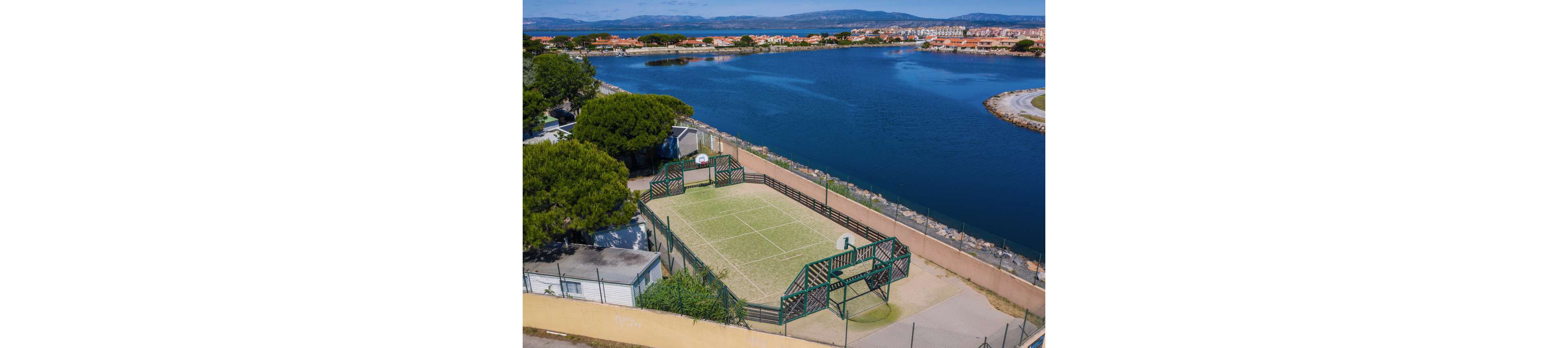 Aerial view of an outdoor multi-sport court situated beside a calm lake. The fenced court features markings for tennis or other ball games and includes basketball hoops at each end. Surrounding the court are trees, pathways, and nearby buildings, with a residential coastal town visible across the water. Distant hills frame the horizon under a clear blue sky.