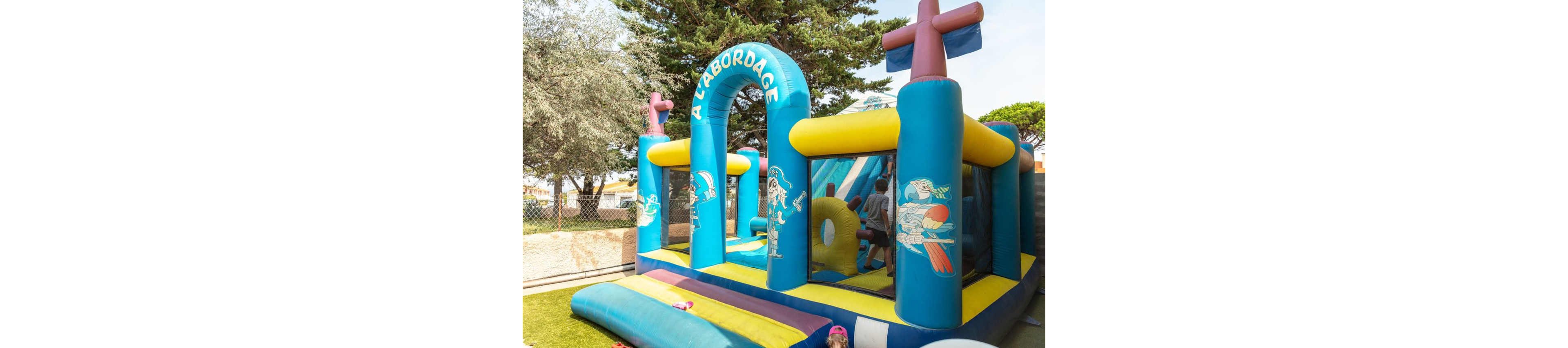 A large pirate-themed inflatable play structure stands in an outdoor area. The blue and yellow bouncy castle features cartoon pirate illustrations and the text "A L'ABORDAGE" over the entrance arch. Children are seen playing inside the structure, climbing and bouncing. Trees, a fence, and nearby buildings frame the sunny recreational setting.