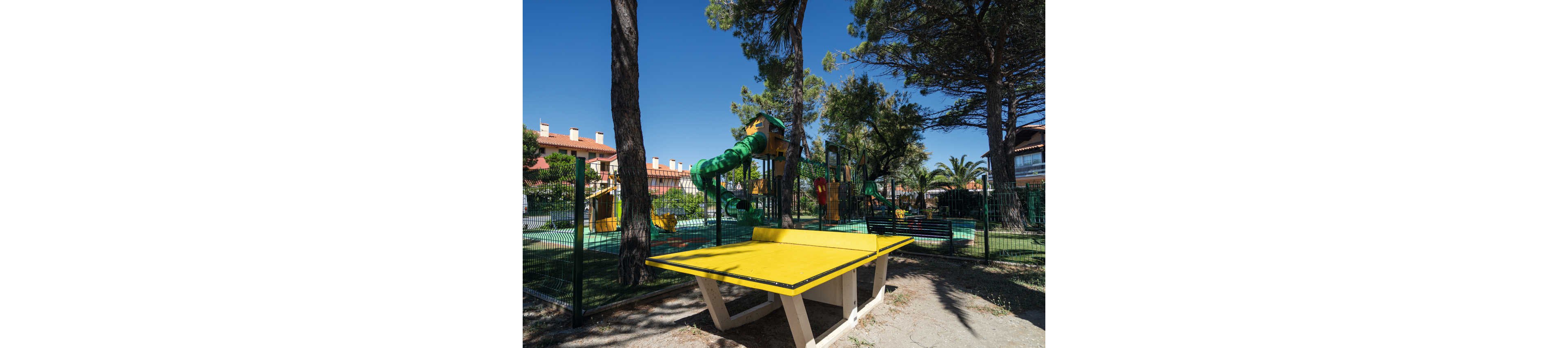 An outdoor recreational area features a bright yellow ping pong table in the foreground. Behind it, a fenced playground includes colorful slides and a green spiral tube slide among tall pine trees. Residential buildings with terracotta roofs are visible in the background under a clear blue sky. The scene suggests a sunny, family-friendly environment in a landscaped park or community space.