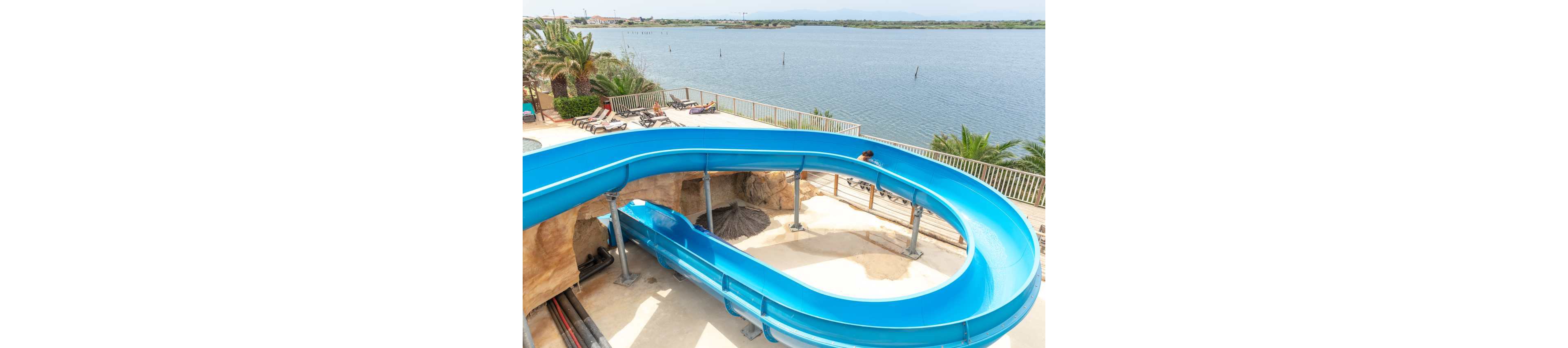 A bright blue waterslide curves around an outdoor recreation area near a large body of water. The slide is elevated on metal supports and winds above a concrete deck and rocky landscaping. Lounge chairs and palm trees sit near the railing, suggesting a resort or water park setting. In the background, calm lake water stretches toward a distant shoreline under a light sky.