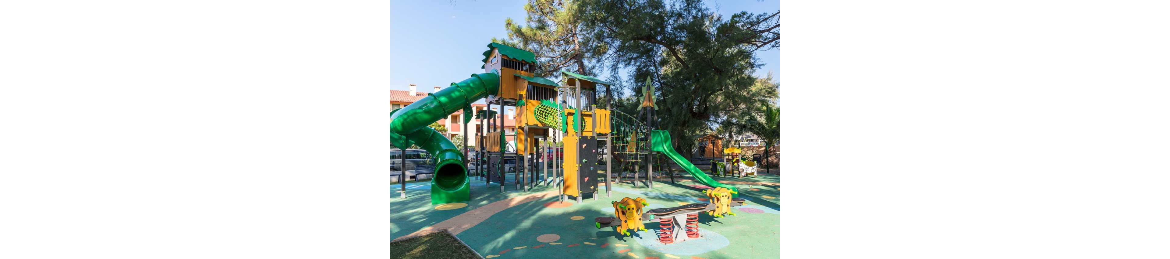 A vibrant outdoor playground features multiple interconnected play structures with towers, bridges, and climbing elements. Bright green tube and open slides dominate the scene, surrounded by cheerful yellow and orange accents. The soft rubberized ground is decorated with playful patterns and animal-themed spring riders. Trees, parked cars, and nearby residential buildings frame the background, indicating a neighborhood park setting.
