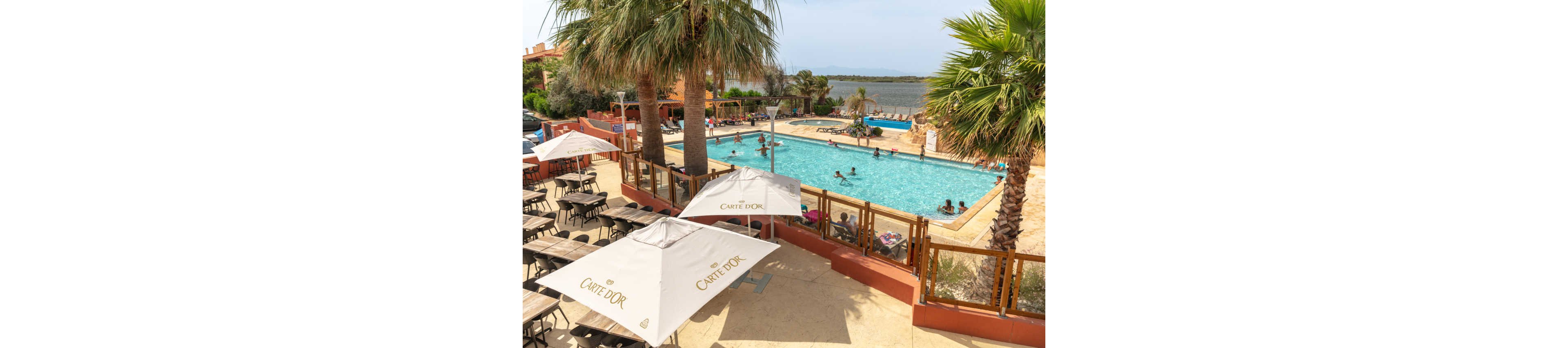 An outdoor resort swimming pool is surrounded by palm trees and sun loungers on a bright day. People of various ages relax and swim in the clear blue water while others sit by the poolside. In the foreground, shaded seating areas with large white umbrellas overlook the pool. Beyond the pool, a calm body of water and distant landscape create a tranquil holiday atmosphere.