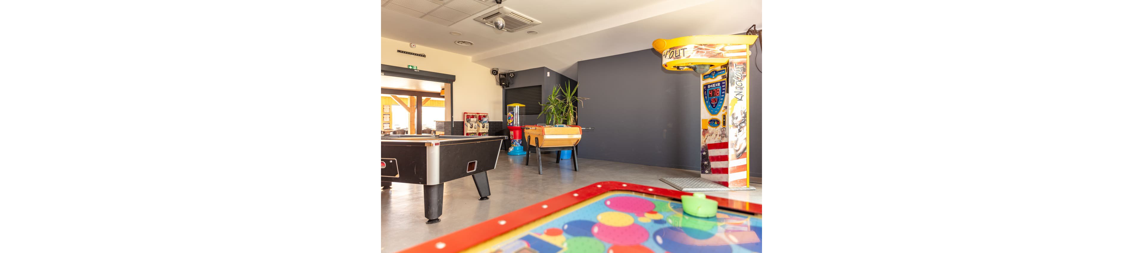 An indoor arcade room features several game tables and machines in a spacious, well-lit setting. A colorful air hockey table is visible in the foreground, with a pool table and foosball table positioned further back. Additional arcade machines and a tall plant line the walls, adding visual interest to the recreational space. Large windows and light-colored walls create an open, inviting atmosphere suitable for leisure and entertainment.